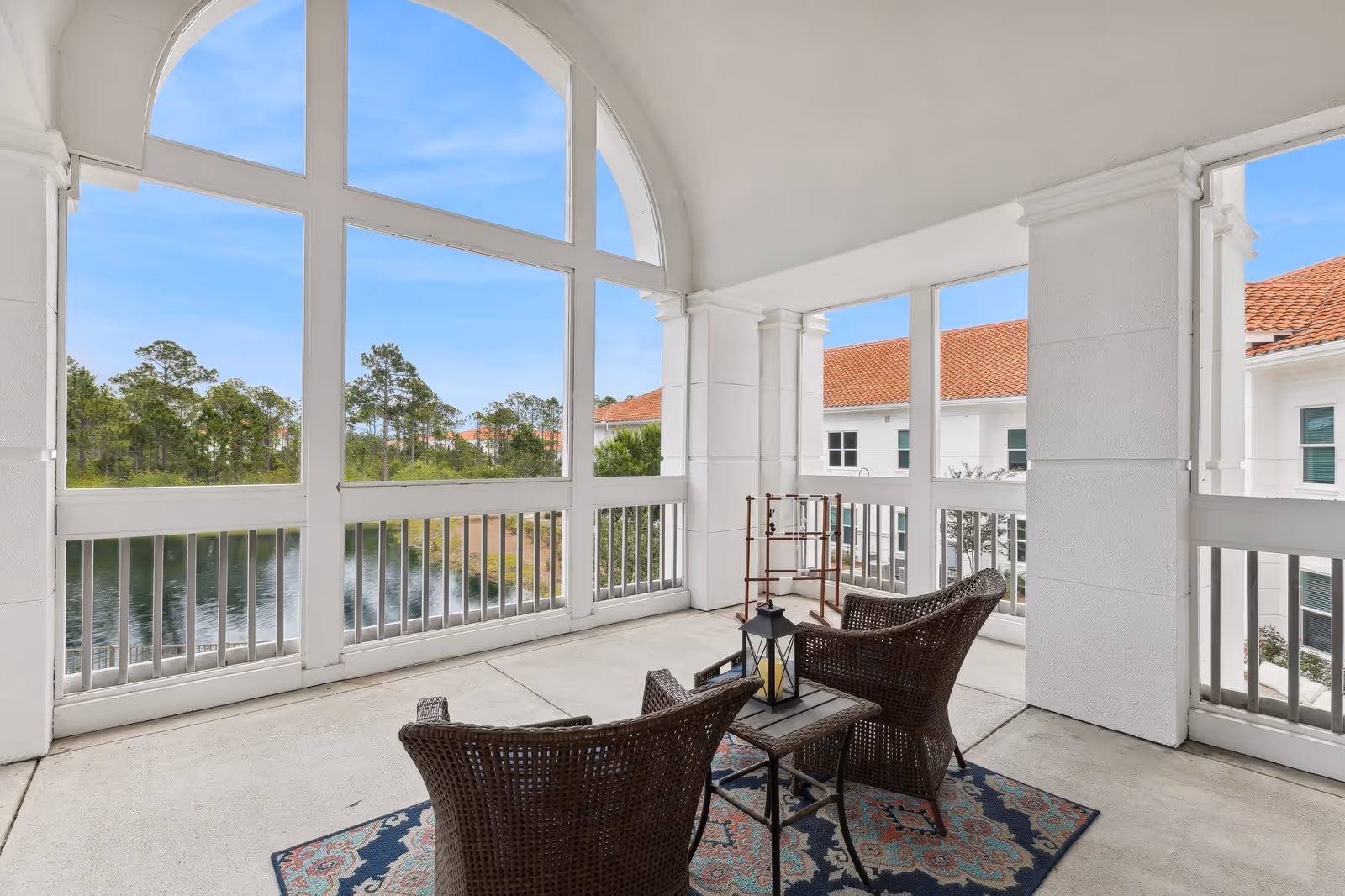 Covered balcony with two wicker chairs, a small table and lantern overlooking a pond and trees through large arched windows.