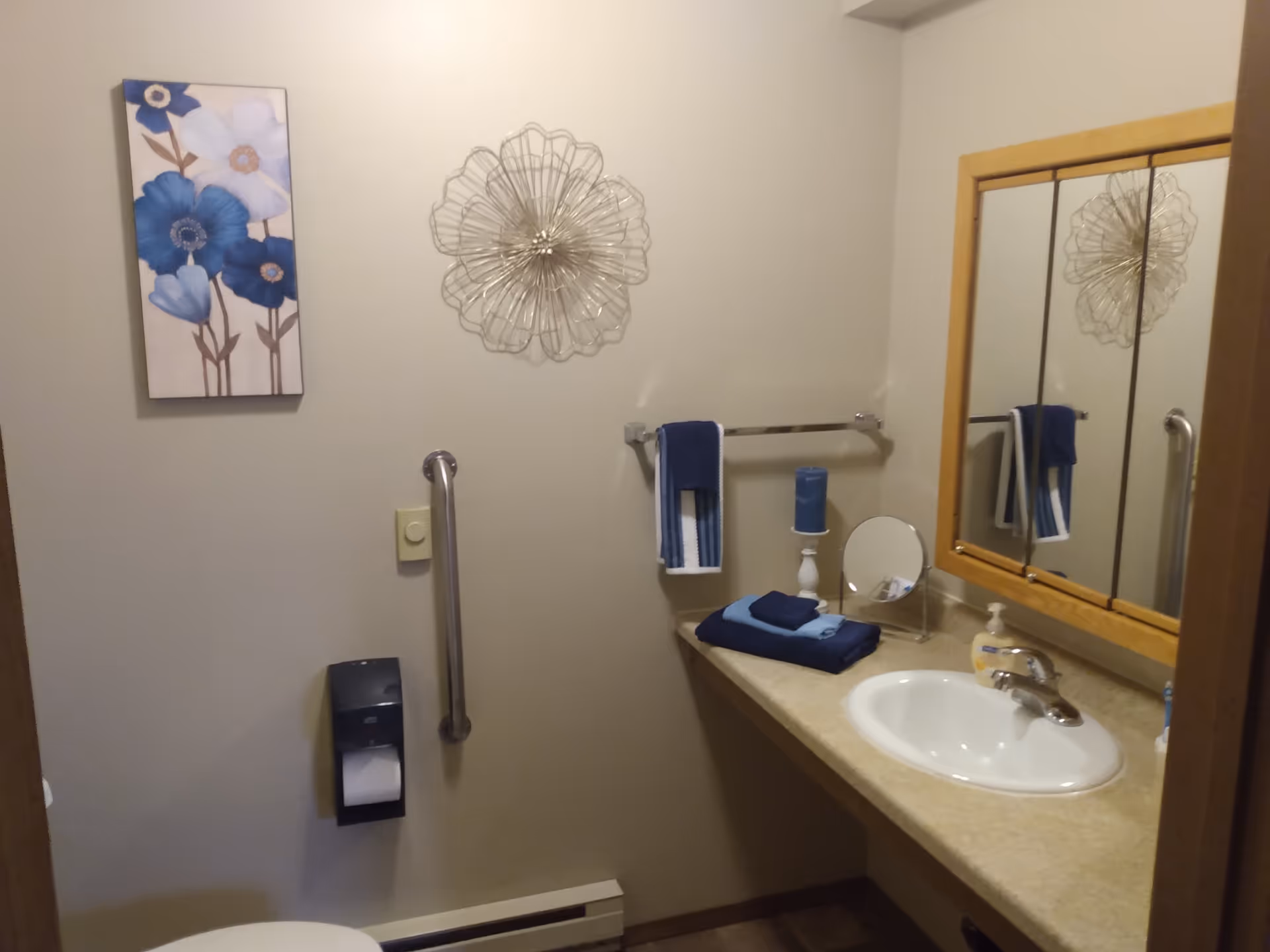 A small bathroom with a beige wall featuring a metal grab bar, a toilet paper holder with toilet paper, a towel rack with a blue and white towel, a countertop with a white sink, folded blue towels, a blue candle on a white stand, a small round mirror, and a soap dispenser. Above the towel rack is a decorative metal wall art piece, and to the left is a framed picture of blue and white flowers.