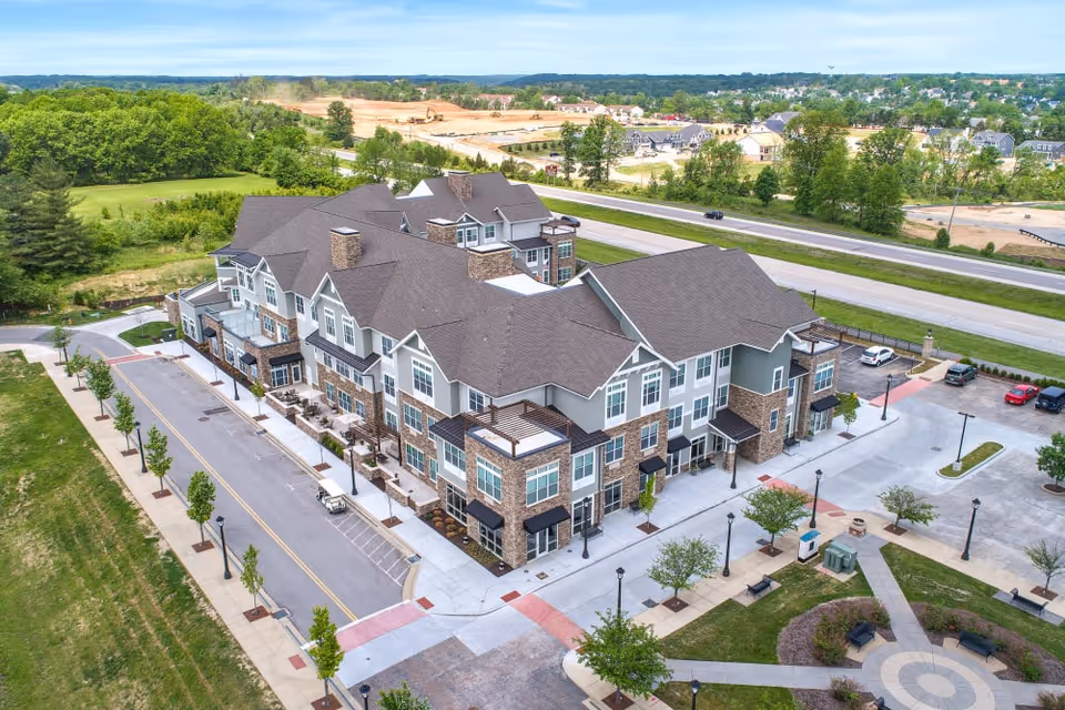 Aerial view of a large, modern senior living facility building with multiple stories, surrounded by parking areas, sidewalks, and landscaped greenery. The building features a combination of stone and siding exterior with numerous windows and small balconies. The surrounding area includes trees, open green spaces, and a road running parallel to the building.