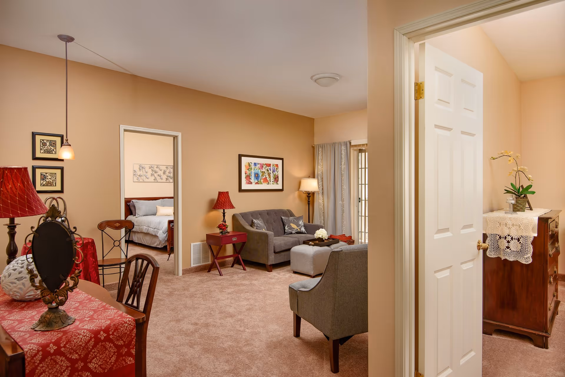 Cozy living room with sofa, armchairs, side tables and lamps, showing a doorway to a bedroom and a dresser.