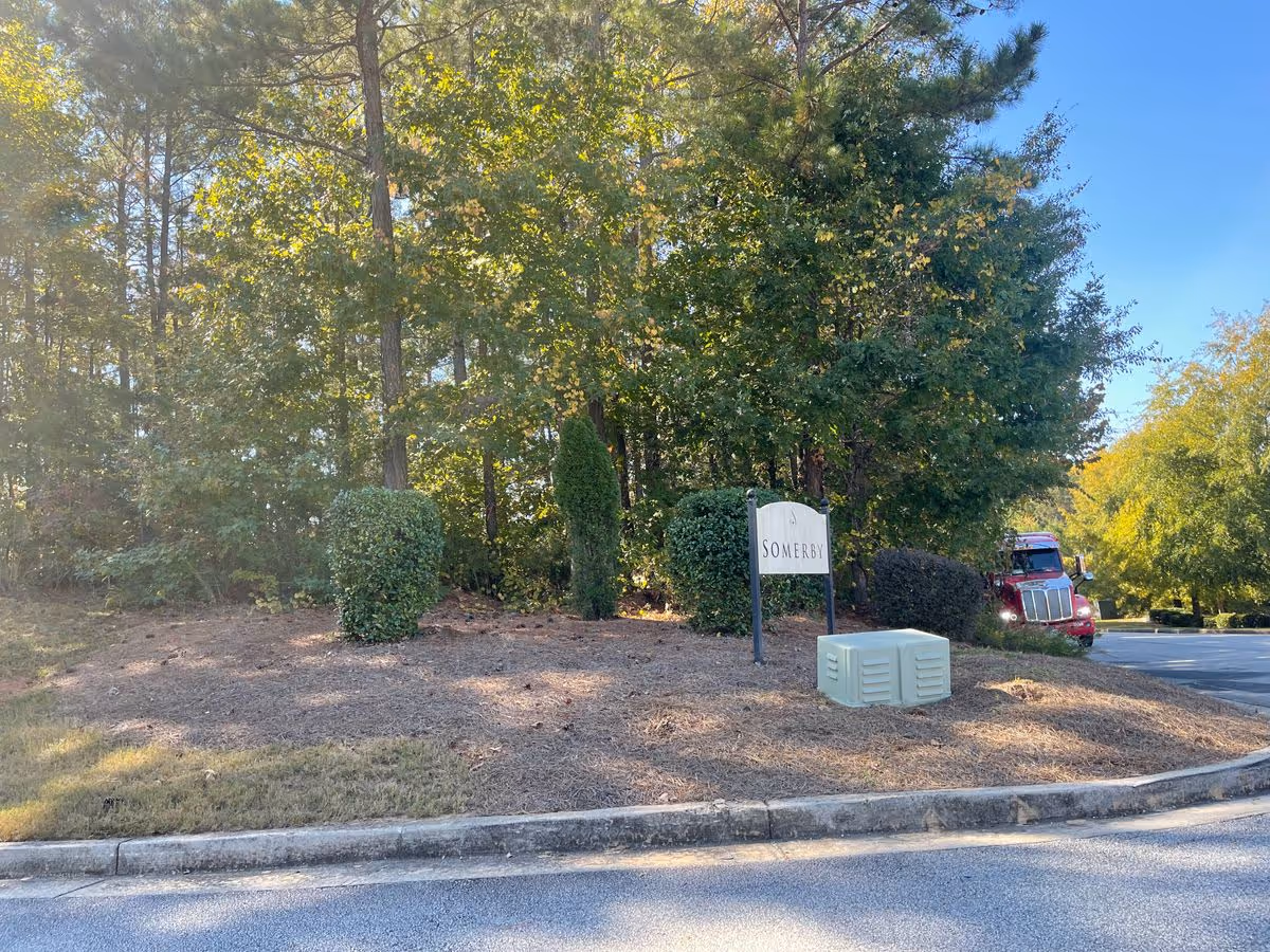 Landscaped roadside entrance with trees, a 'SOMERBY' sign, a utility box, and a red truck partially visible.