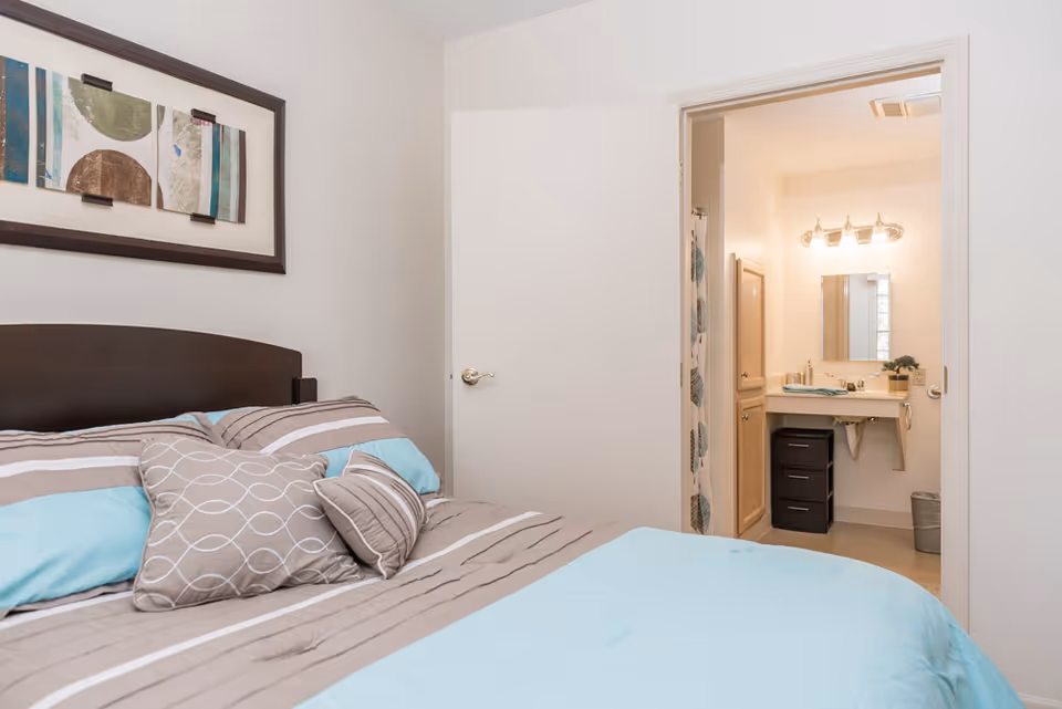 A bedroom with a bed made up with blue and beige striped bedding and multiple pillows. Above the bed is a framed abstract artwork. To the right, an open door reveals a bathroom with a sink, mirror, light fixture, and a shower curtain with a leaf pattern.