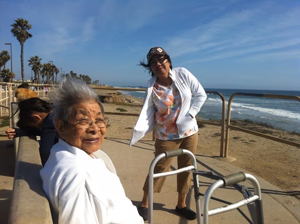 An elderly woman with gray hair and glasses sits on a bench near the beach, smiling at the camera. Another woman stands nearby wearing a white jacket, floral shirt, and cap, with a walker in front of her. The ocean and a sandy beach with palm trees are visible in the background under a clear blue sky.