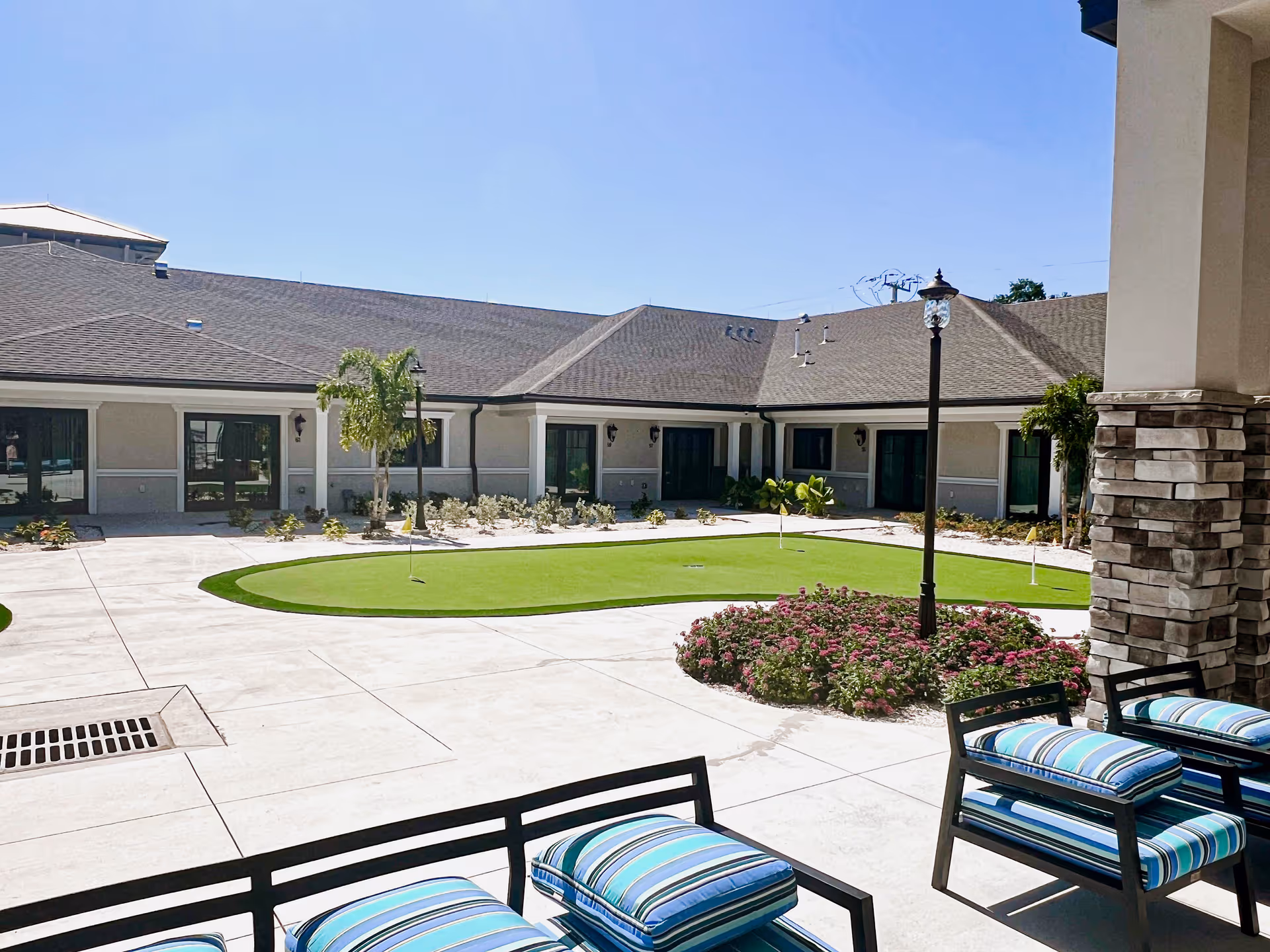 Outdoor courtyard area at Villas of Holly Brook in Sarasota, Florida, featuring a putting green, landscaped flower beds, palm trees, and patio seating with striped cushions under a clear blue sky.