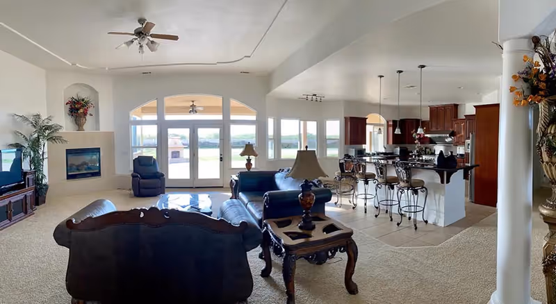 Spacious open-plan living room and kitchen with leather sofas, a fireplace, bar stools at an island, and large windows/doors overlooking the outdoors.