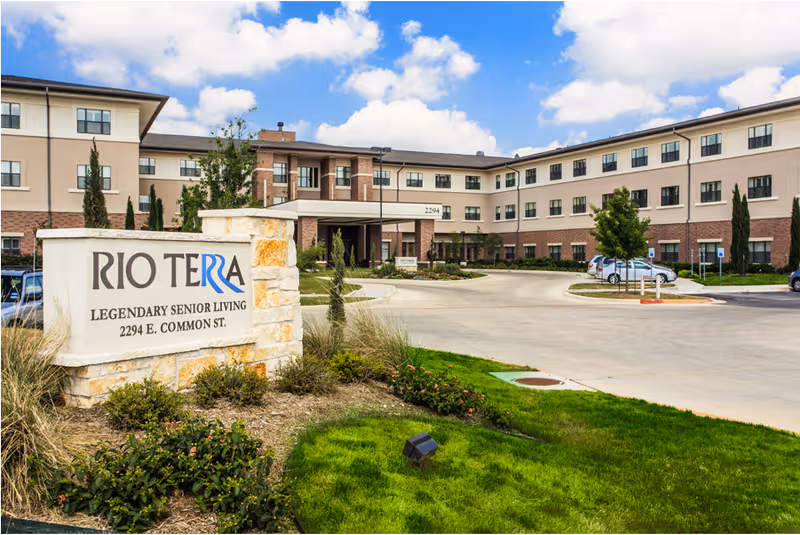Front exterior of the Rio Terra senior living building with landscaped grounds and a stone sign in the foreground.