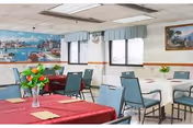 Interior view of a dining room with several tables covered with tablecloths in red and white. Each table has a small vase with flowers. The room has blue chairs, windows with valances, and a large mural on one wall depicting a harbor scene with boats and buildings.