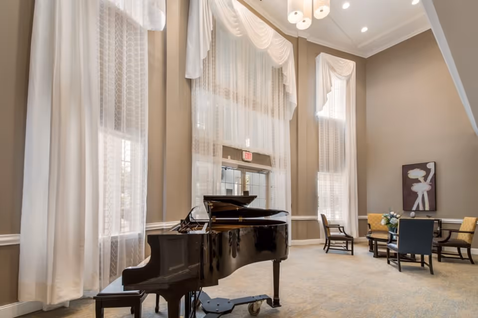 Grand piano in a high-ceilinged lounge with tall draped windows and a seating area.