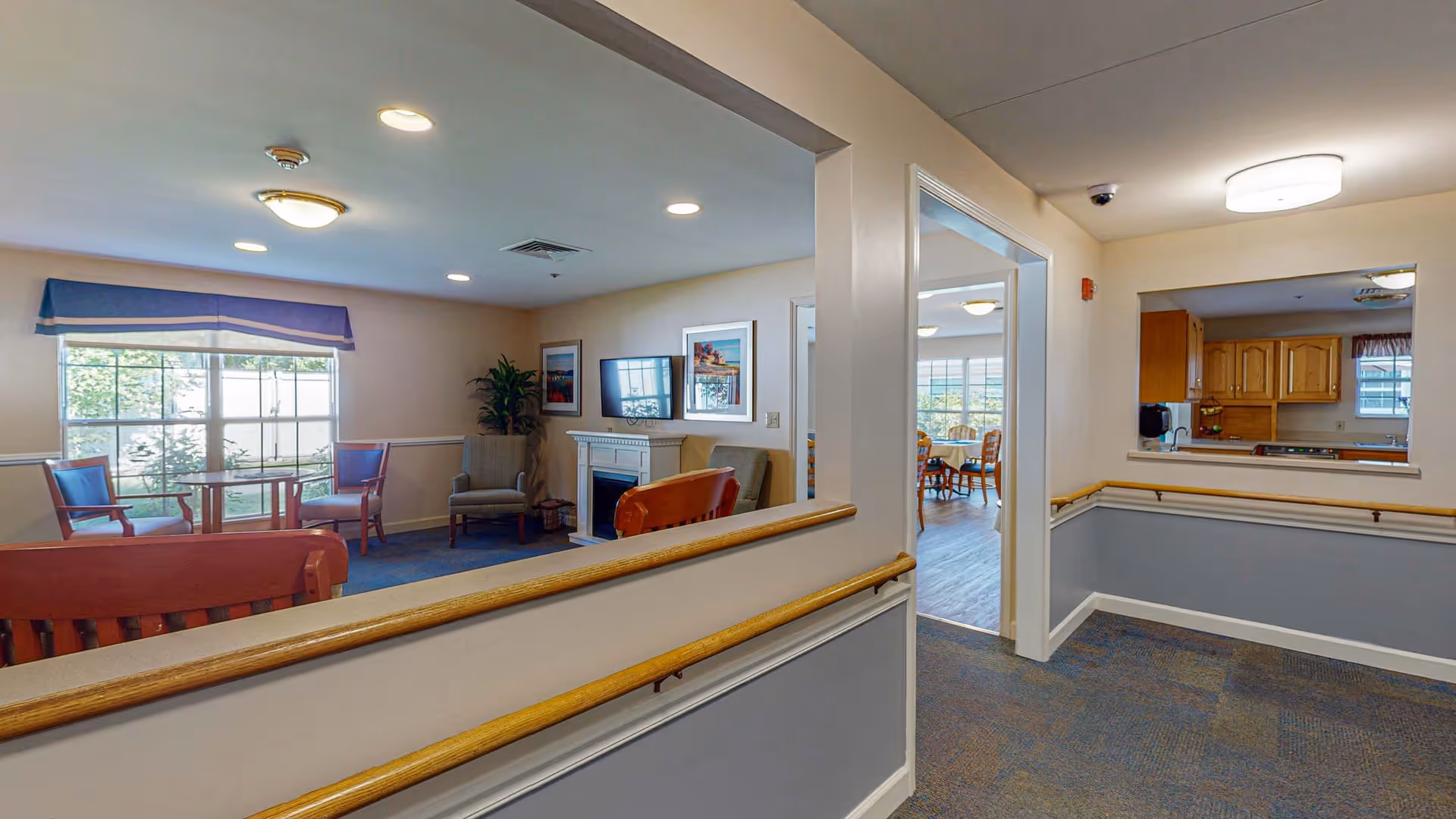 Interior view of a senior living facility showing a common area with chairs, a small table, a fireplace, and framed pictures on the wall. There is a hallway with handrails leading to a dining area with tables and chairs, and a kitchen window opening to the hallway. The space is well-lit with ceiling lights and natural light from windows.