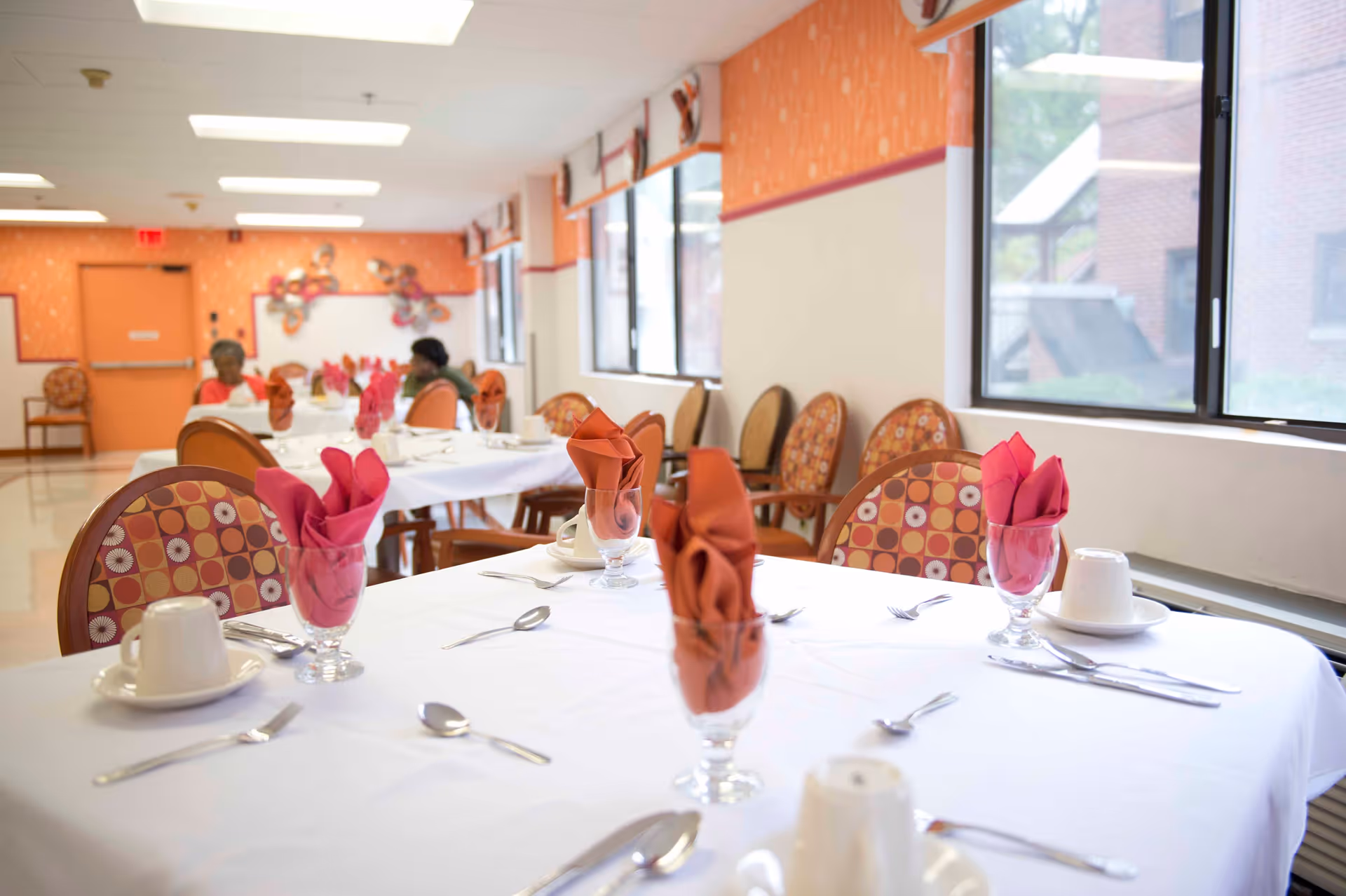 A dining room in a senior living facility with tables covered in white tablecloths, each set with cups, saucers, silverware, and glasses holding folded red and orange napkins. The room has patterned chairs, large windows letting in natural light, and orange walls with decorative accents. Two elderly individuals are seated at a table in the background.