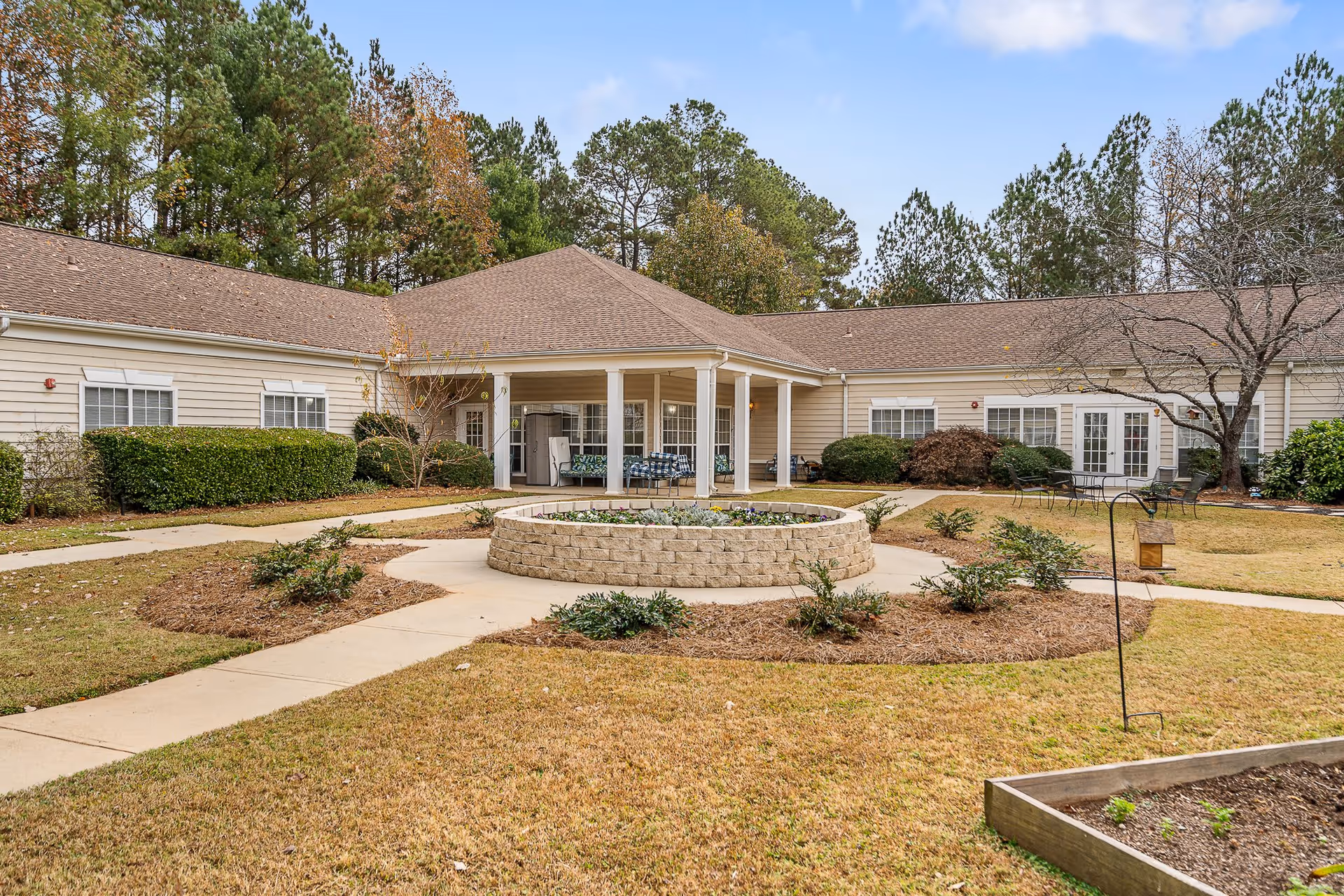 Outdoor courtyard area of a senior living facility with a circular raised flower bed in the center, surrounded by paved walkways, grass, and small shrubs. The building has beige siding, white trim, and a covered patio with seating. Trees and bushes are visible around the perimeter under a partly cloudy sky.