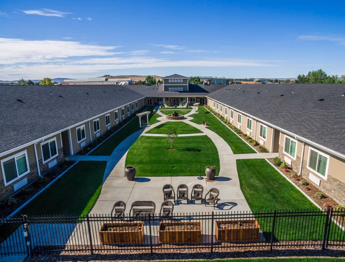 Aerial view of a U-shaped single-story building surrounding a well-maintained courtyard with green grass, paved walkways, outdoor seating, and decorative planters under a clear blue sky.