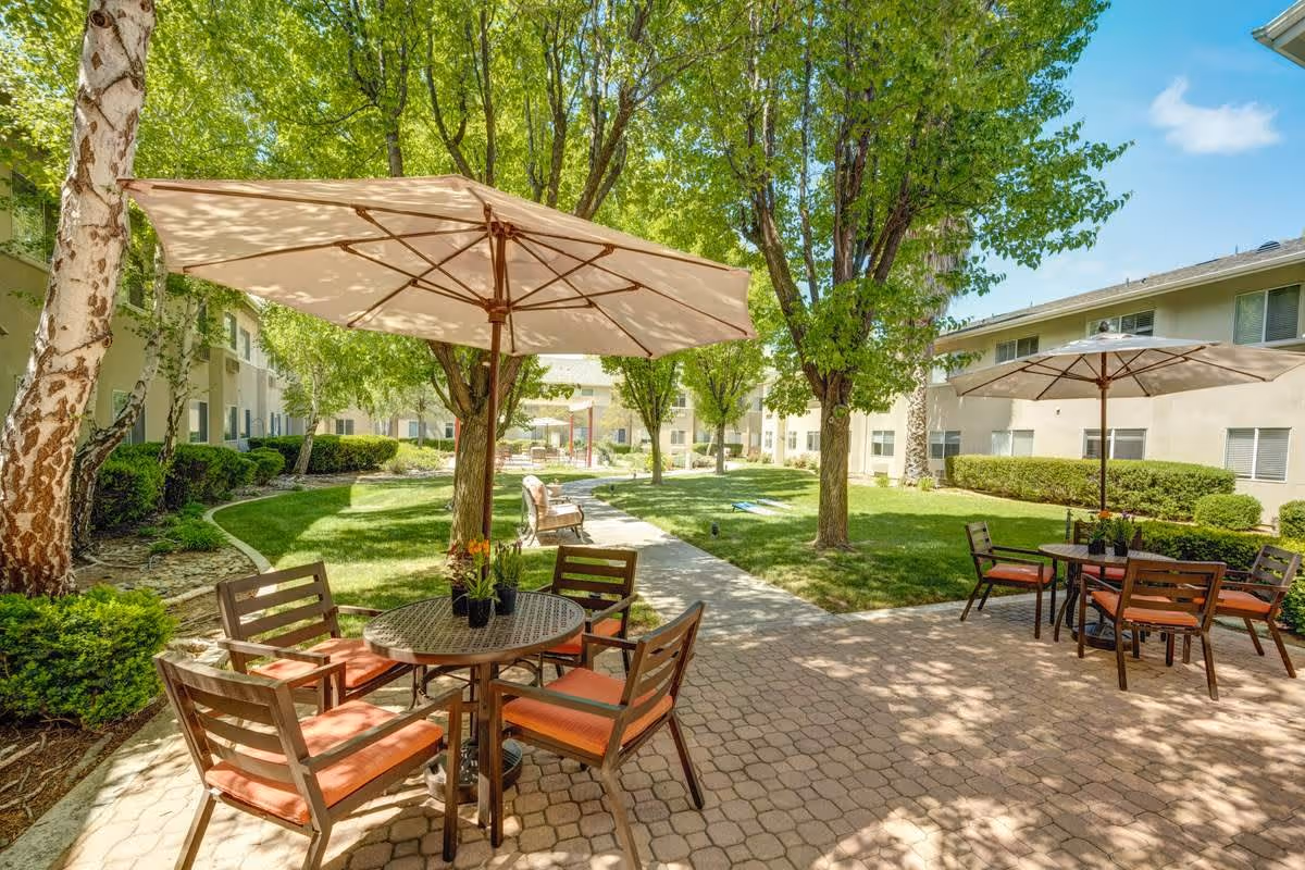 Outdoor patio area at Creston Village with round tables and chairs featuring orange cushions, shaded by large beige umbrellas. The patio is surrounded by green grass, trees, and bushes with a paved walkway running through the middle. Residential buildings are visible in the background under a clear blue sky.