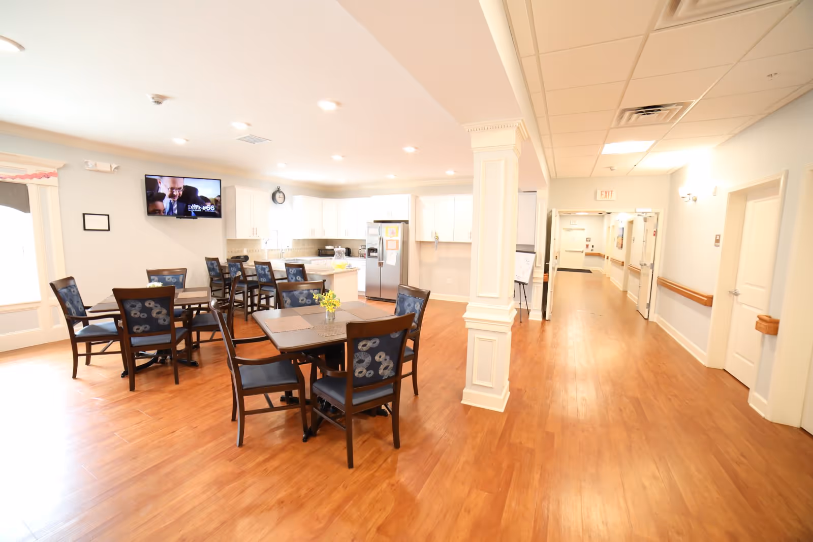 A bright and spacious dining area in a senior living facility with wooden floors, several tables and chairs with blue patterned cushions, a kitchen area with white cabinets and stainless steel refrigerator in the background, and a hallway leading to other rooms.