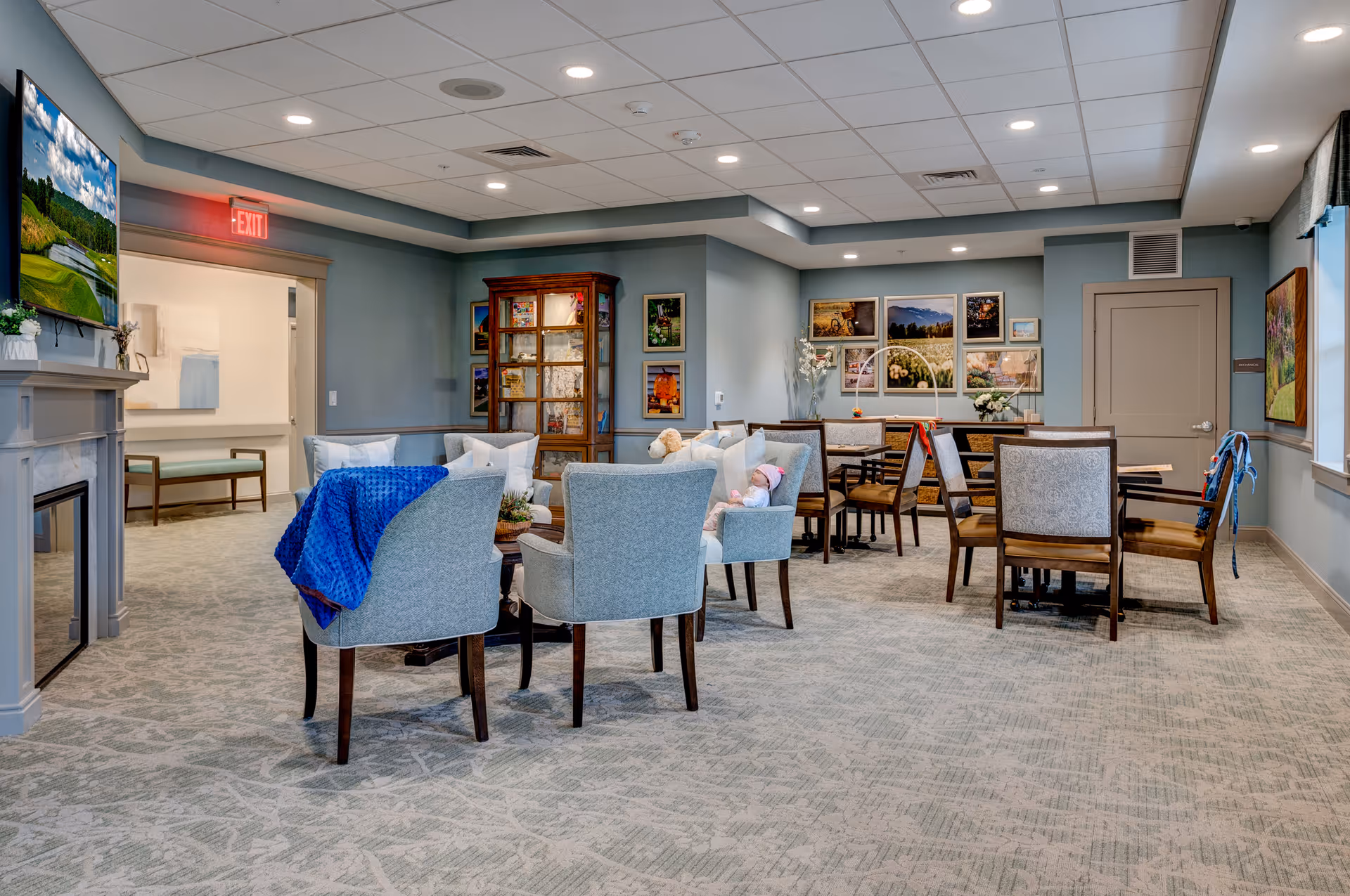 A cozy senior living common area with light blue walls and carpeted floor. The room features several upholstered chairs arranged around a small table, a fireplace with a TV mounted above it, and a wooden cabinet with glass doors. There are framed pictures on the walls and a dining table with chairs in the background. A blue blanket is draped over one chair, and a doll is seated on another chair. The room is well-lit with recessed ceiling lights.