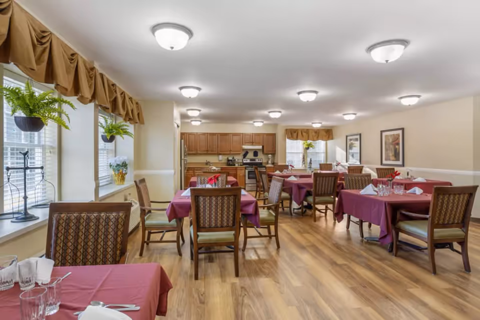 A dining room in a senior living facility with several tables covered in burgundy tablecloths, each set with glasses, napkins, and silverware. Wooden chairs with patterned upholstery surround the tables. The room has wood flooring, beige walls with white trim, and multiple ceiling lights. There are windows with brown valances and hanging plants, and a kitchen area with wooden cabinets and stainless steel appliances is visible in the background.