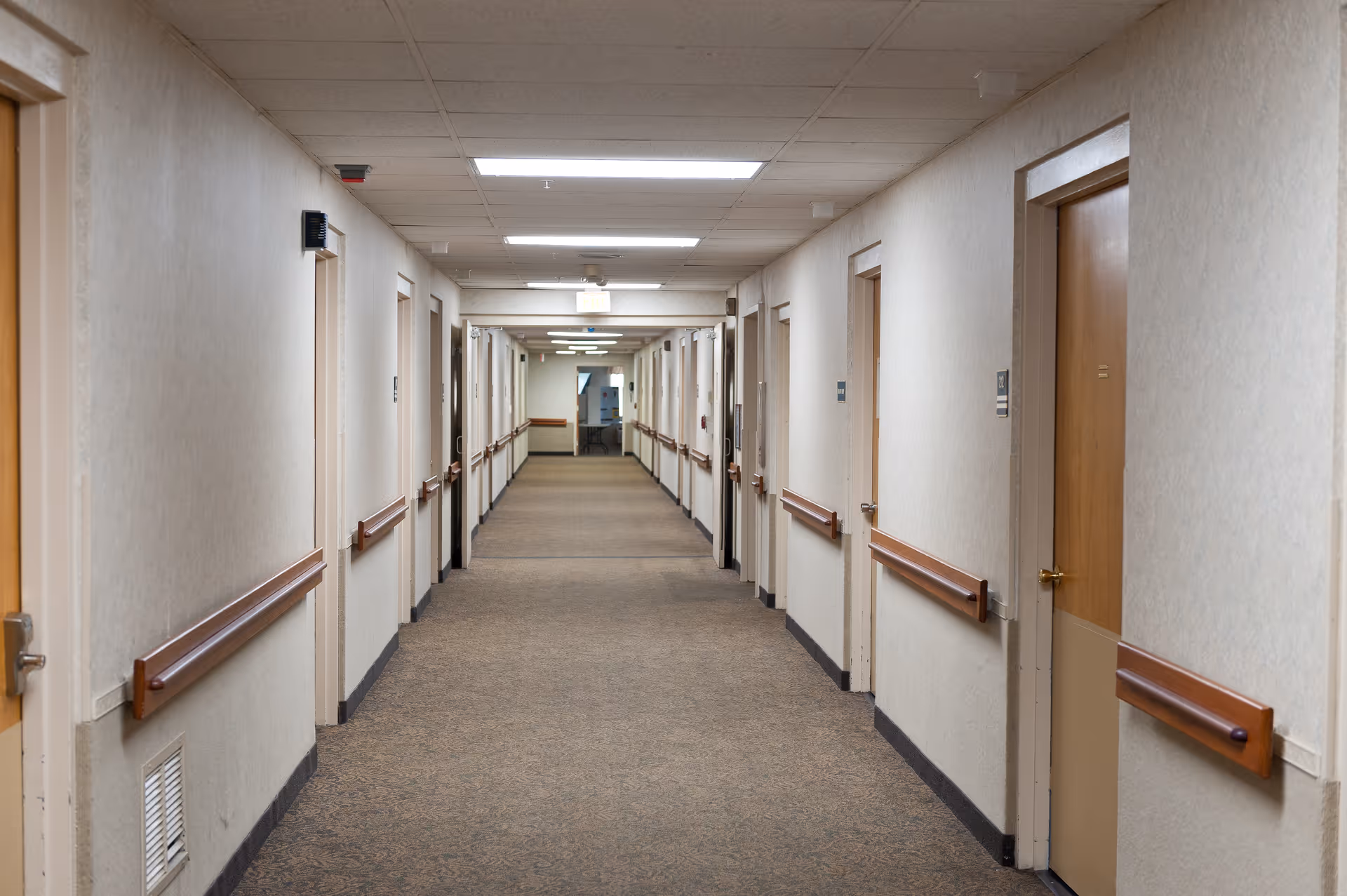 A long, empty hallway in a healthcare facility with beige walls, brown handrails on both sides, multiple closed wooden doors, and fluorescent ceiling lights.