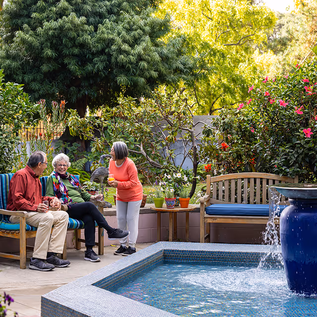 Three older adults chatting on benches beside a tiled fountain in a lush garden patio.