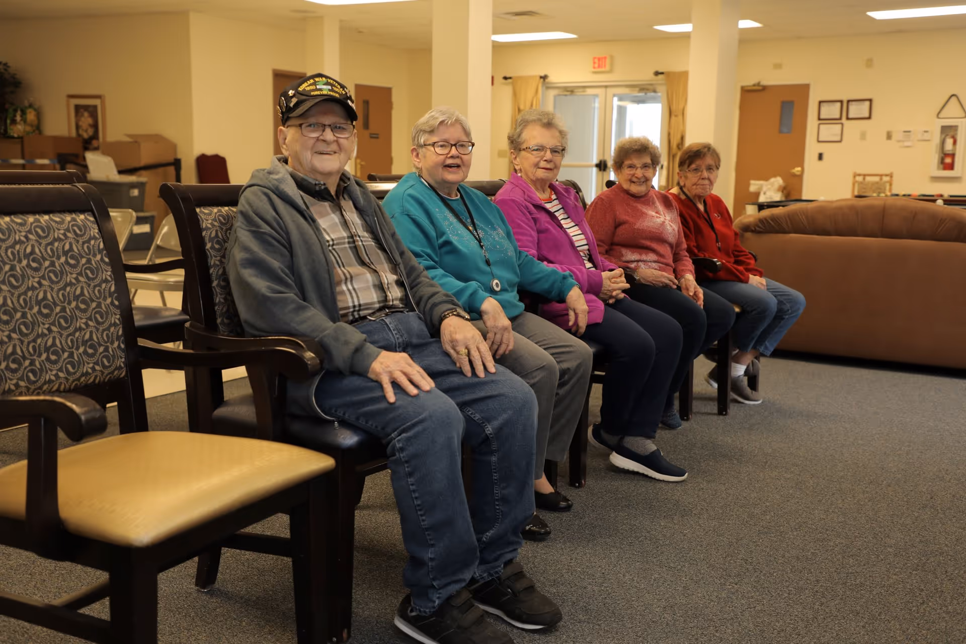 Five elderly individuals sitting in a row on chairs in a common area of a senior living facility. The room has beige walls, carpeted floor, and some furniture including a brown couch and additional chairs. There is a door with an exit sign in the background.