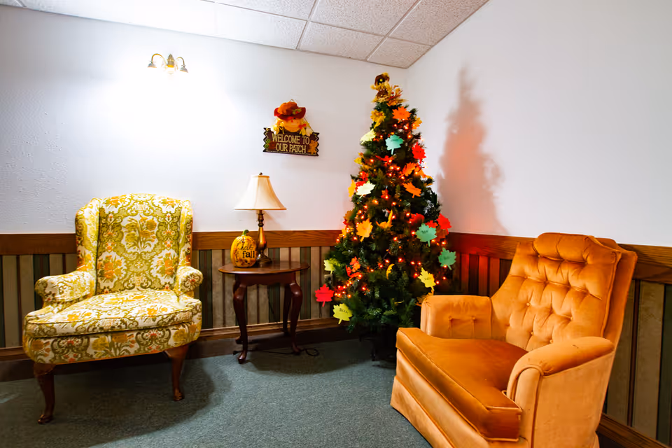 A cozy corner in a senior living facility featuring two vintage armchairs, one with a floral pattern and the other in orange upholstery, separated by a small wooden side table with a lamp and a decorative pumpkin. A decorated Christmas tree with colorful paper leaves and lights stands in the corner, and a wall sign above reads 'Welcome to Our Patch.'