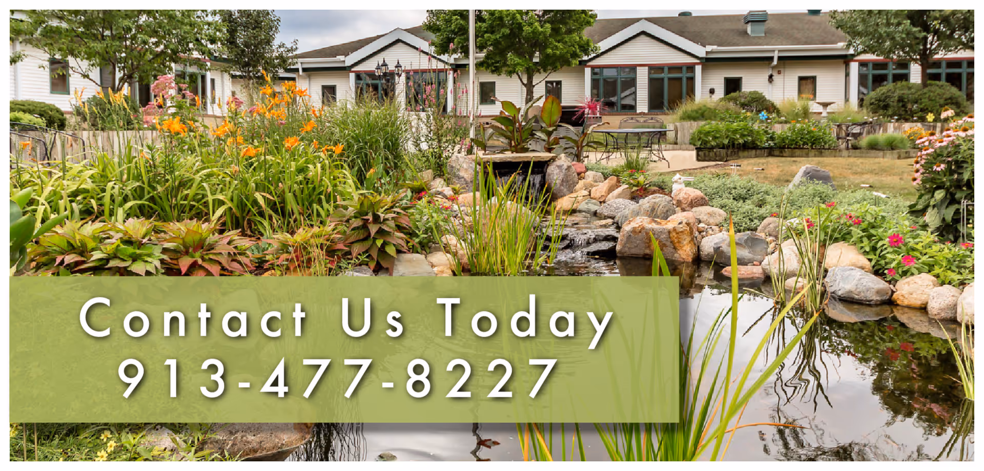 A landscaped garden area with a small pond surrounded by rocks and various plants and flowers. In the background, there are single-story buildings with large windows and outdoor seating areas. The sky is partly cloudy.