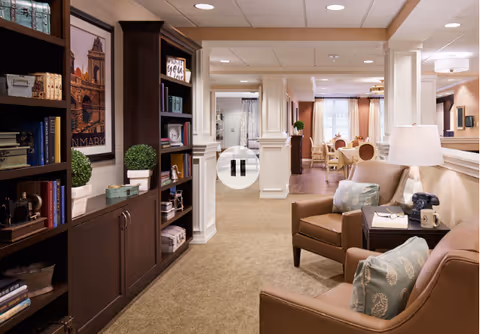 Cozy lounge in a senior living facility with beige seating, lamps, and bookshelves.