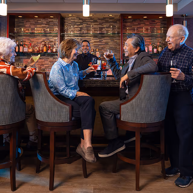 Several older adults seated at a bar in a well-lit lounge, smiling and raising glasses in a toast.