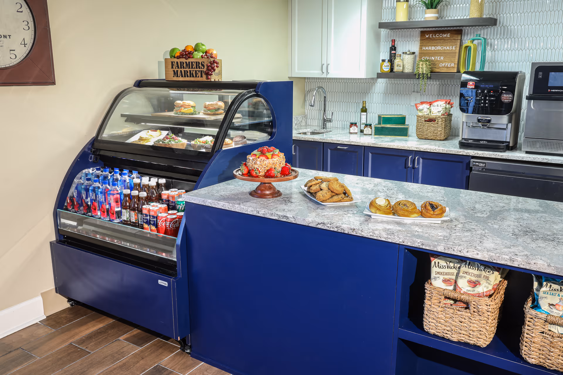 A kitchen or snack counter area with a blue cabinet and marble countertop. On the counter are plates with cookies, pastries, and a cake topped with strawberries. Behind the counter is a refrigerated display case with bottled drinks and sandwiches. The background features a sink, shelves with bottles and plants, a coffee machine, and a sign that reads 'WELCOME HARBORCHASE COUNTER OFFER'.