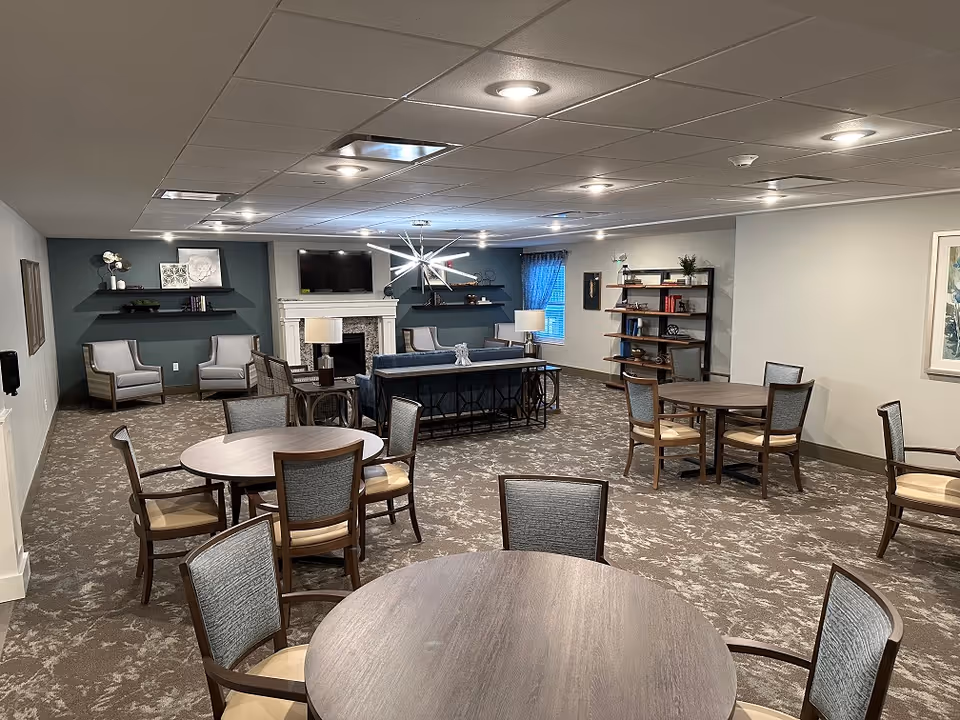 A spacious senior living common area with multiple round tables and chairs arranged on a patterned carpet. The room features a seating area with armchairs and a sofa facing a fireplace with a mounted TV above it. Shelves with decorative items and books are on the walls, and the ceiling has recessed lighting.