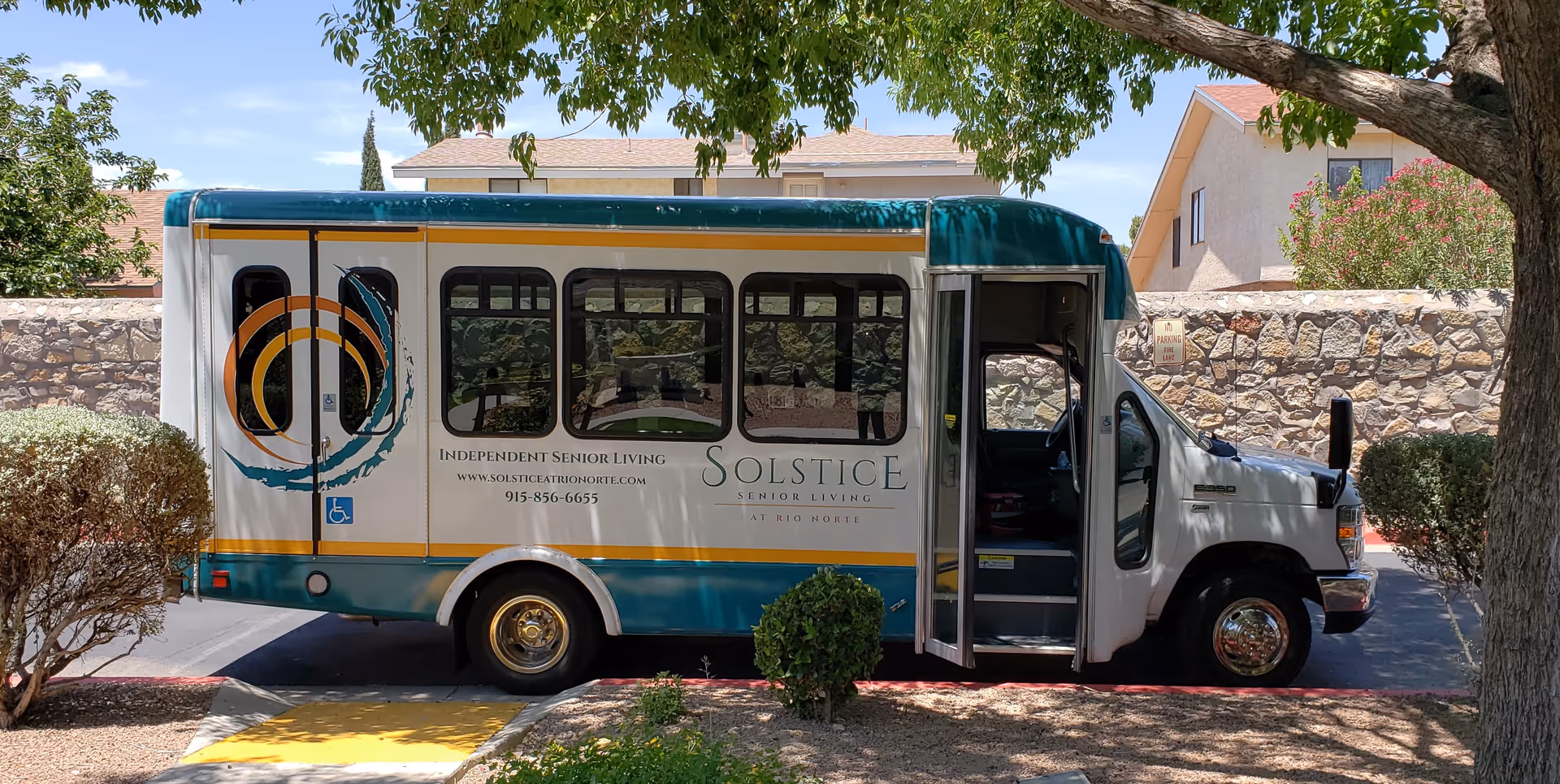 A Solstice Senior Living shuttle bus parked under trees with its passenger door open in front of a stone wall.