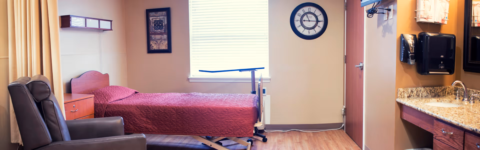 A nursing facility patient room with a single bed covered in a red quilt, a recliner, bedside table, wall clock, and a sink/vanity area.