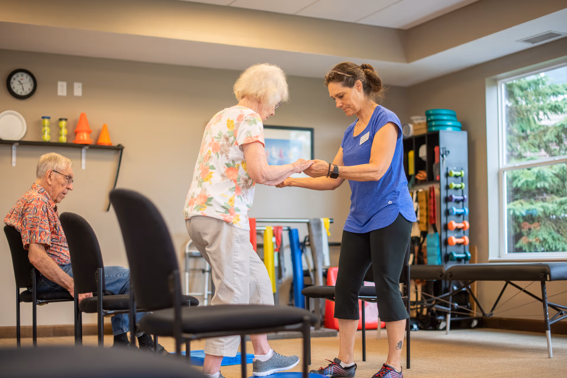 A senior woman is assisted by a staff member in a blue shirt as she practices standing or walking in a therapy or activity room. An elderly man sits on a chair nearby watching. The room has exercise equipment, chairs, and large windows with a view of trees outside.