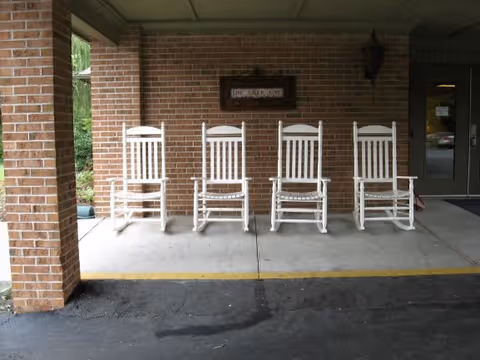 Four white wooden rocking chairs lined up on a covered porch area with a brick wall background and a sign mounted on the wall. There is a doorway to the right and a pillar to the left.