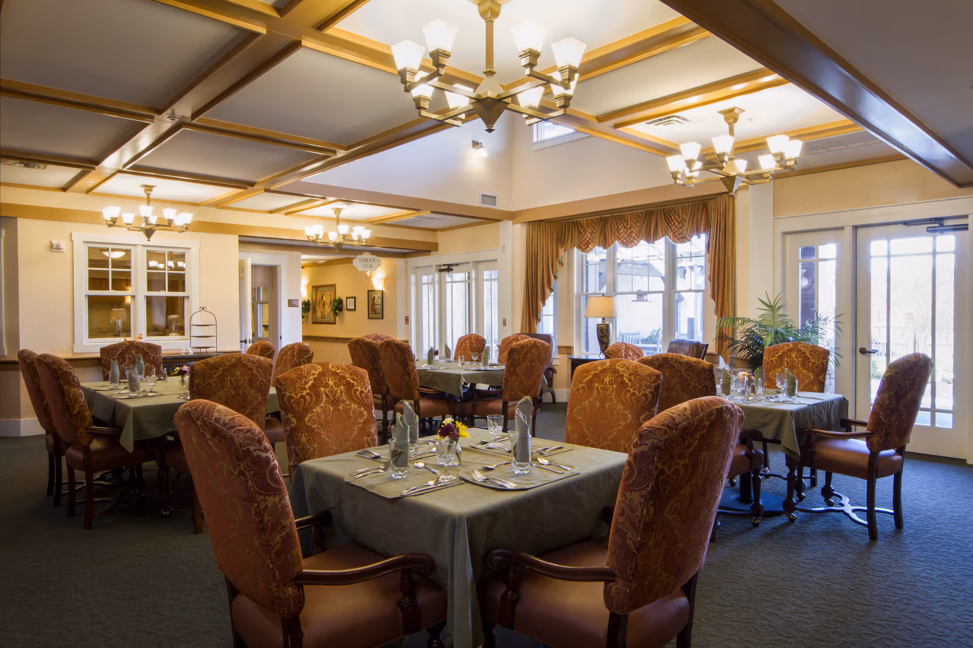 A well-lit dining room with several tables covered in green tablecloths, each set with glasses, napkins, and silverware. The chairs have ornate red and gold upholstery. Large windows with patterned curtains allow natural light to fill the room. Elegant chandeliers hang from the ceiling, and there is a door leading outside.