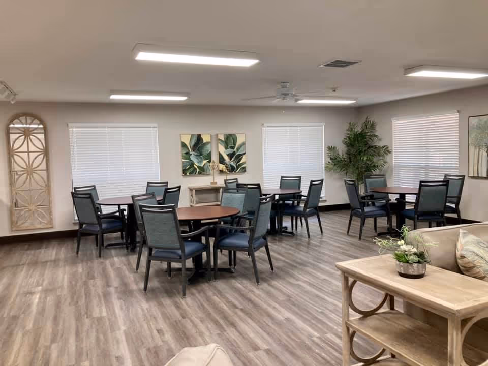 A bright and clean dining area with multiple round tables and chairs arranged neatly. The room has wood-look flooring, white walls, and several windows with blinds. There are decorative plants and framed artwork on the walls, along with a beige couch and a wooden side table with a small plant in the foreground.