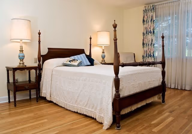 A neatly made four-poster wooden bed with a white textured bedspread and decorative pillows in a bedroom with hardwood floors. There are two bedside tables, each with a lamp, and a window with sheer curtains and floral drapes.