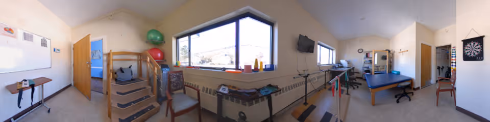 A wide panoramic view of a rehabilitation therapy room with exercise equipment including steps, therapy balls, parallel bars, and a treatment table. The room has large windows letting in natural light, a dartboard on the wall, a whiteboard, and some chairs and desks.