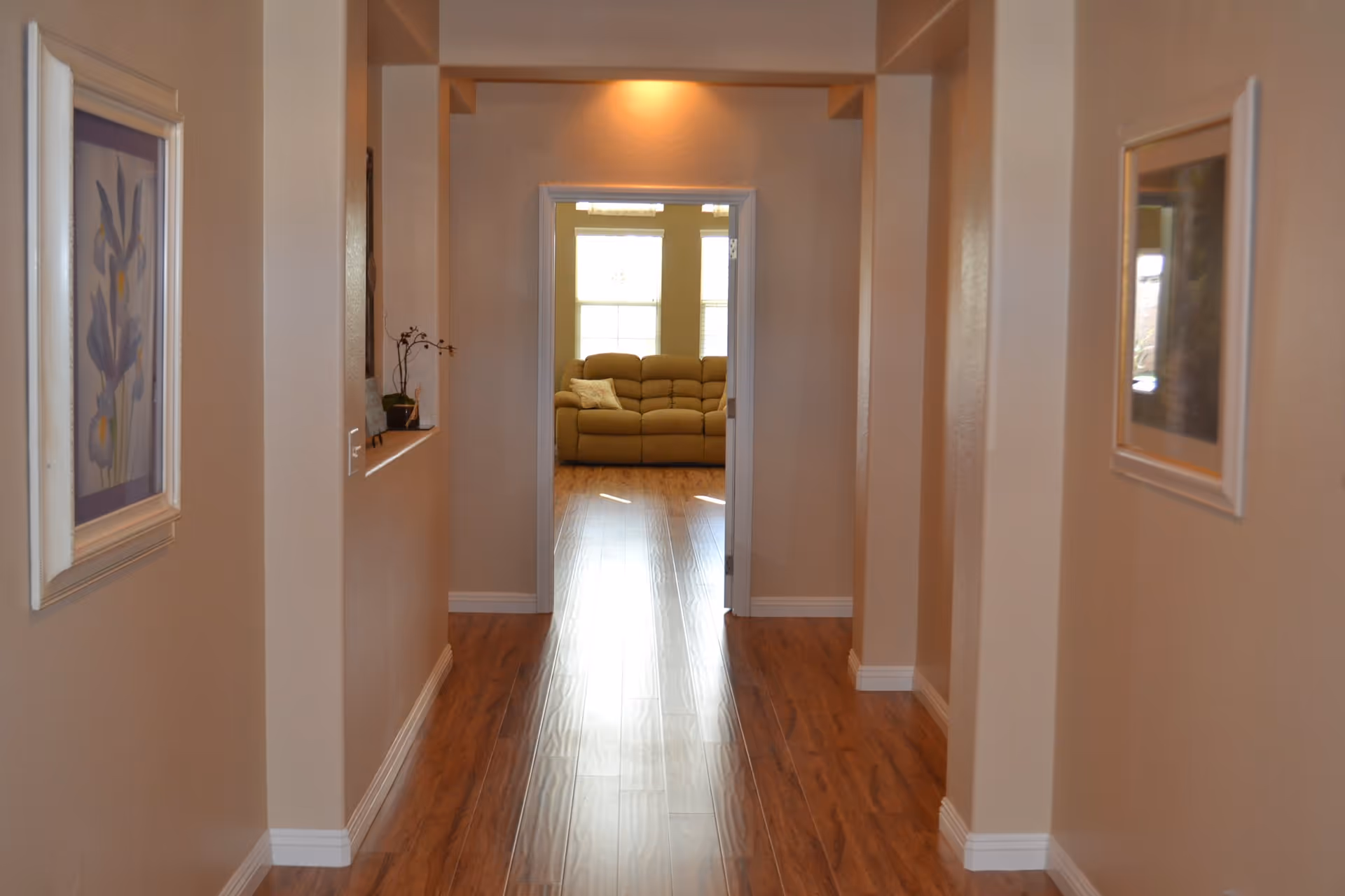 A hallway with wooden flooring and beige walls, decorated with framed artwork on both sides. At the end of the hallway, there is a doorway leading to a room with a beige sofa and large windows letting in natural light.