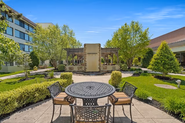 Outdoor seating area in a garden with a round metal table and four chairs with cushions, surrounded by green bushes and trees. In the background, there is a pergola structure and a multi-story building under a clear blue sky.