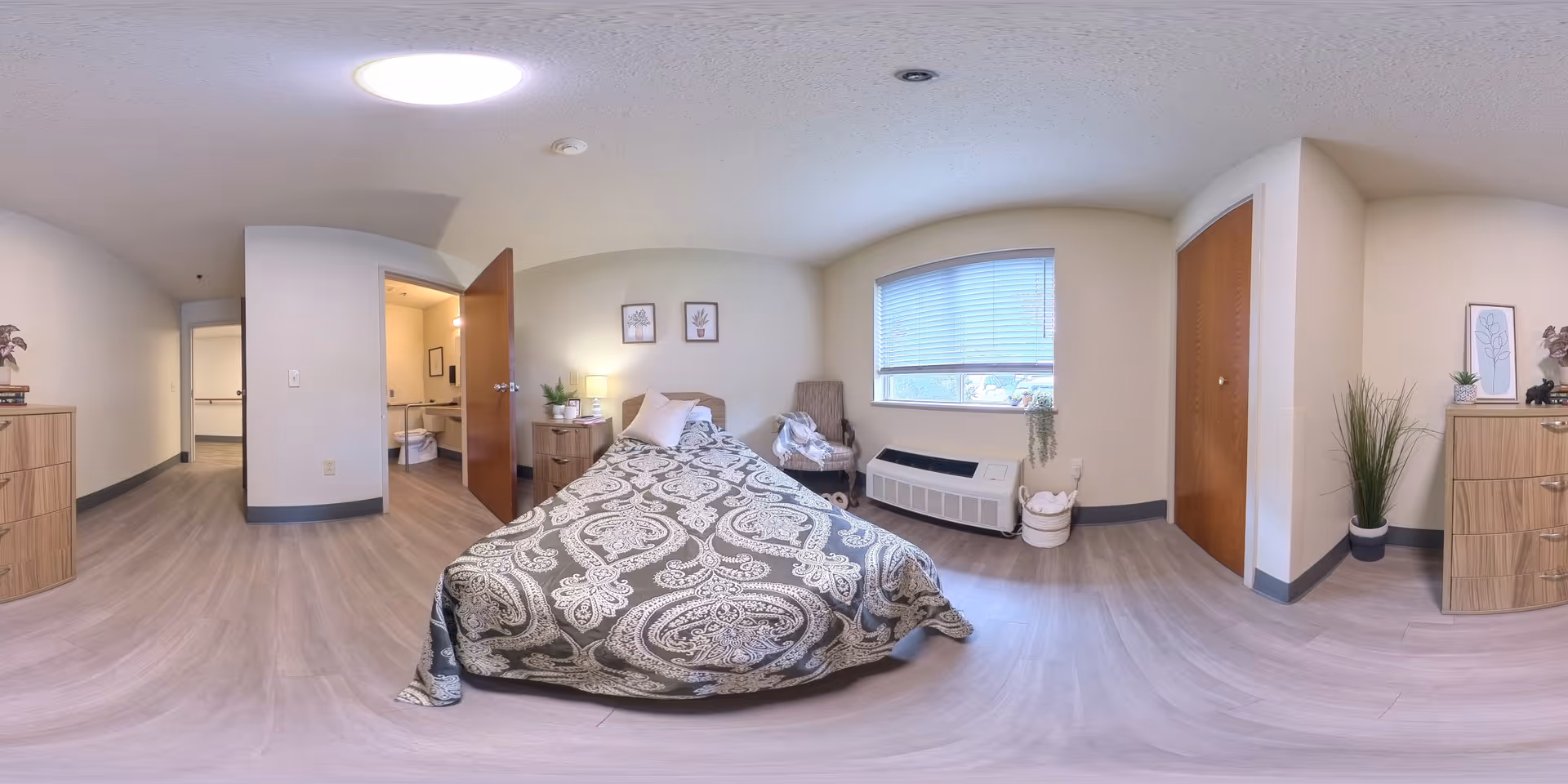 A senior living facility bedroom with a single bed covered in a patterned bedspread, a wooden nightstand with a lamp and plant, a chair near a window with blinds, a heating unit below the window, and two wooden dressers with decorative plants and framed artwork on the walls. An open door reveals a bathroom with a sink and toilet.