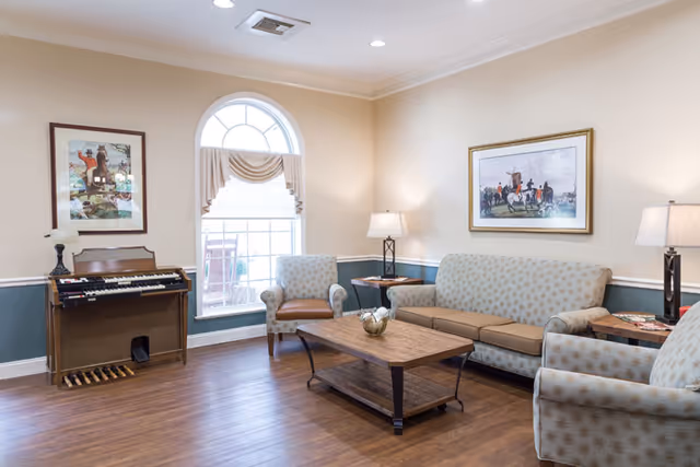 A cozy living room with tan walls, arched window, sofas, chairs, coffee table, and piano.