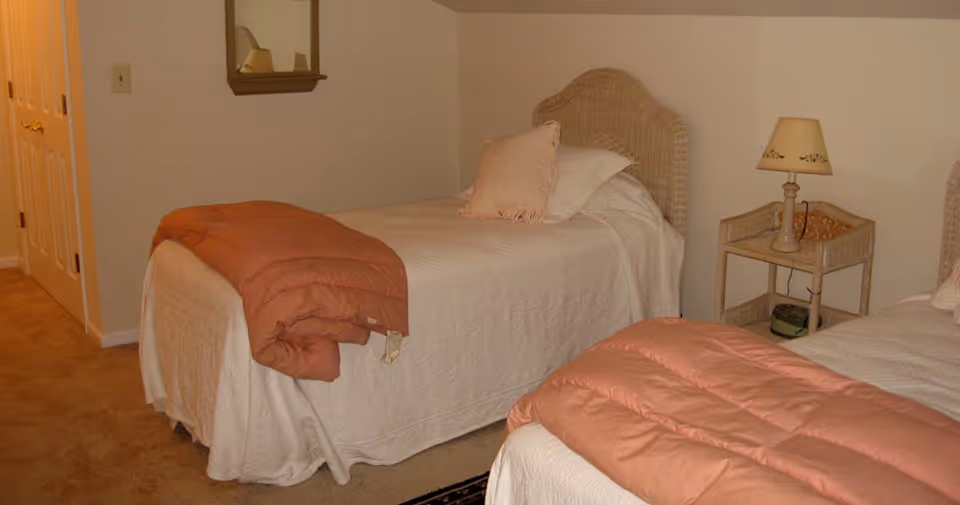 A bedroom with two single beds, each covered with white bedspreads and pinkish-orange comforters folded at the foot. One bed has a pink pillow and a white pillow. Between the beds is a small wicker nightstand with a lamp on top. The room has beige carpet and light-colored walls with a mirror hanging above one bed.