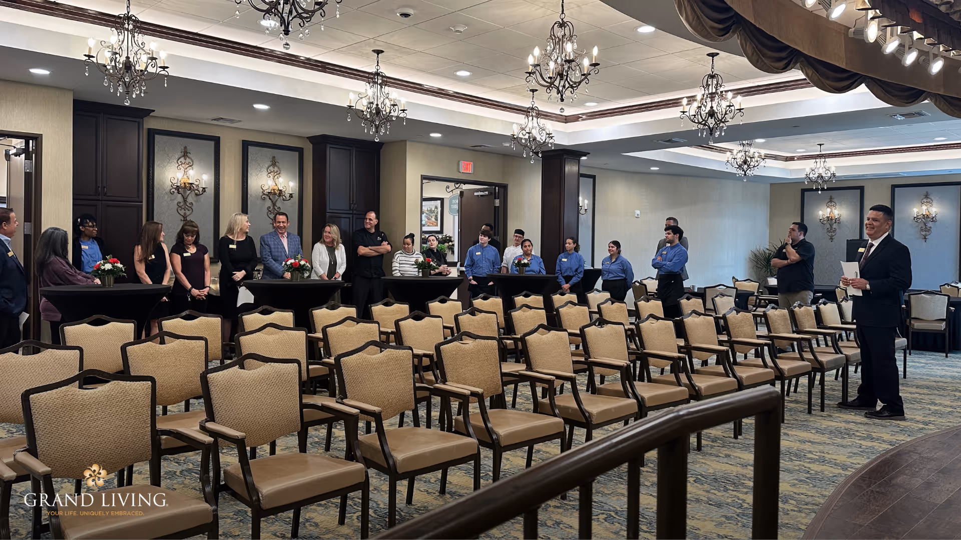 A decorated community event room with rows of chairs and staff lined up at the front while a man in a suit speaks.