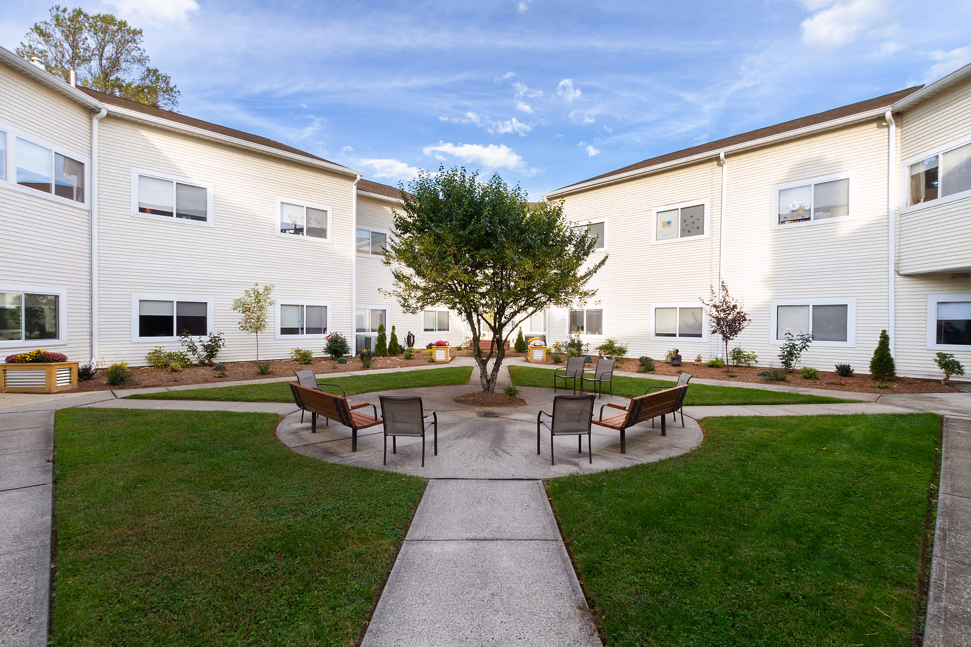 Outdoor courtyard area of a senior living facility with a circular concrete seating area featuring benches and chairs around a tree. The courtyard is surrounded by a two-story white building with multiple windows and landscaped with grass, small trees, and shrubs under a partly cloudy sky.