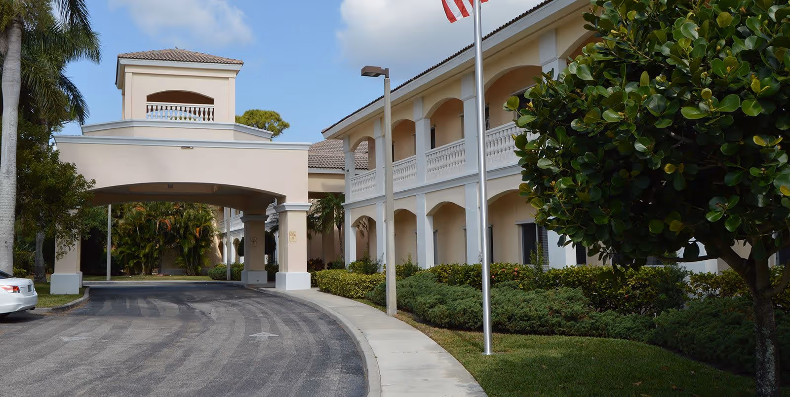 Exterior view of Courtyard Gardens of Jupiter senior living facility showing a two-story building with arches, a covered driveway entrance, palm trees, bushes, and an American flag on a flagpole.
