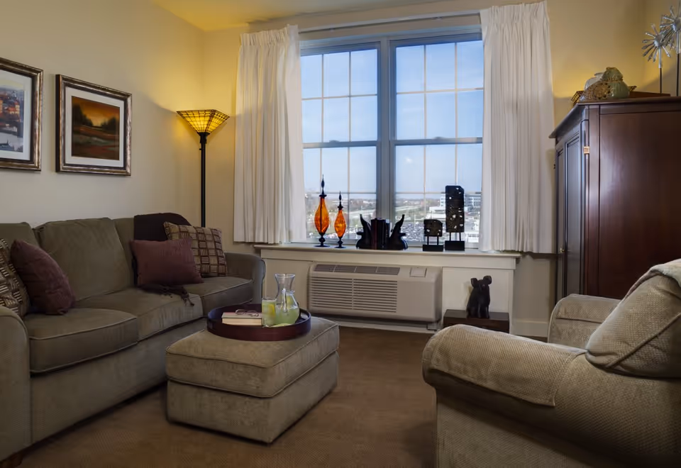 Cozy living room with a sofa, armchair and ottoman centered around a tray, a large window with decorative glass vases, floor lamp and wooden cabinet.