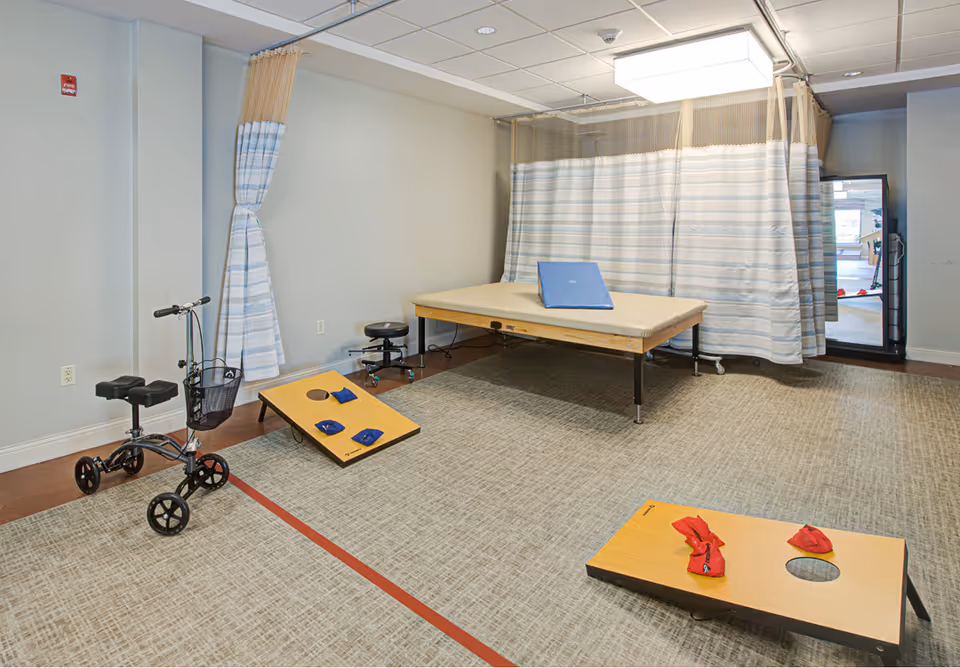 A rehabilitation room with a padded therapy table, two bean bag toss boards with bean bags, a knee scooter, a stool, and a privacy curtain. The room has a carpeted floor and a large mirror on the right side.