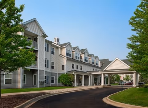 Exterior view of a multi-story senior living facility building with beige siding, multiple windows, and a covered entrance driveway. The building is surrounded by green trees and a clear blue sky.