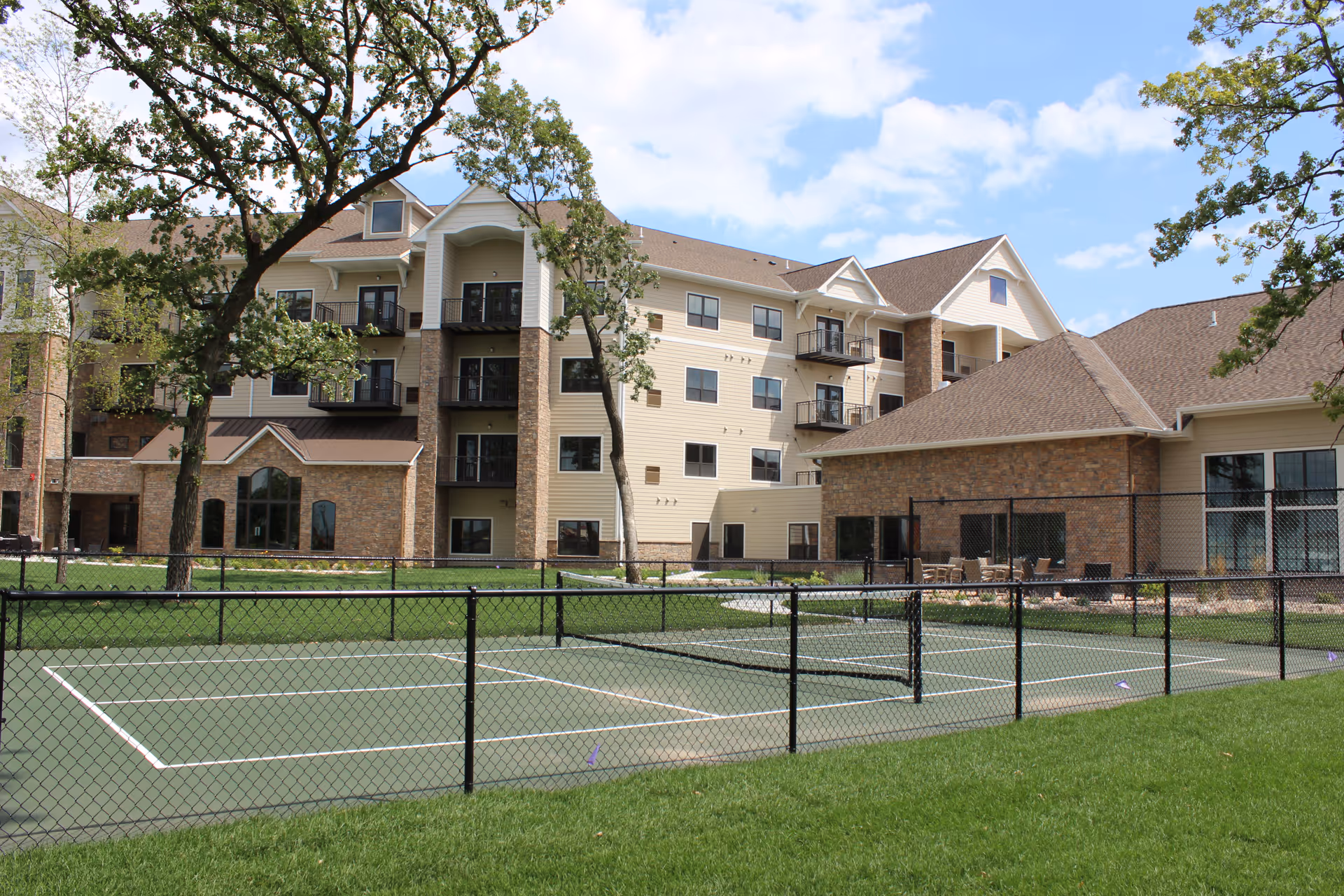 Exterior of a multi-story senior living building with a fenced tennis court and lawn in the foreground.