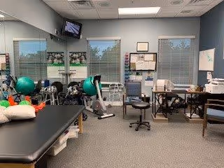 Interior view of a therapy or rehabilitation room with exercise equipment, therapy balls, chairs, a treatment table, and various medical supplies. The room has large windows with blinds, a wall-mounted TV, and a bulletin board with papers pinned on it.